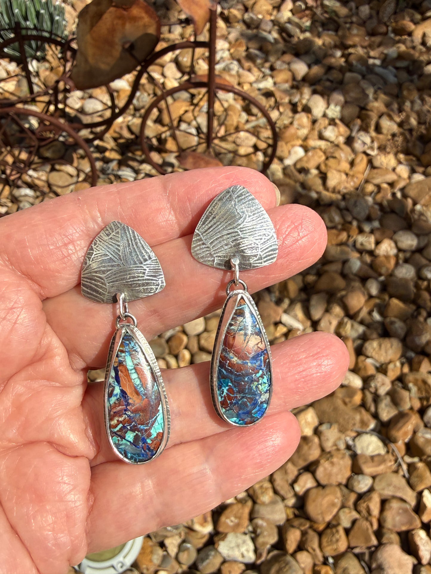 Bluebird azurite and Argentium silver drop earrings held in hand, showing textured triangle stud tops and teardrop bezel-set stones with vivid blue, teal, copper, and purple matrix