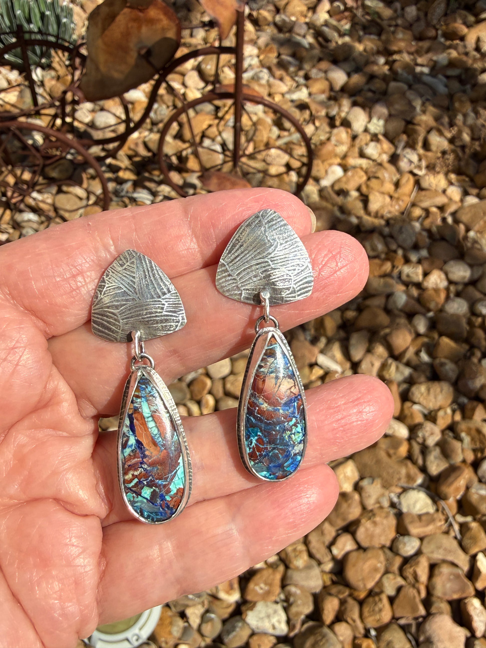 Bluebird azurite and Argentium silver drop earrings held in hand, showing textured triangle stud tops and teardrop bezel-set stones with vivid blue, teal, copper, and purple matrix