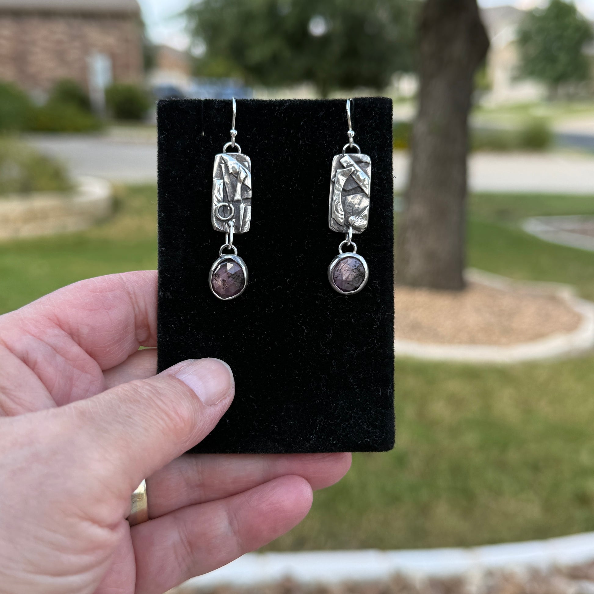 Pink sheen sapphire and patchwork silver dangle earrings on black velvet display card outdoors