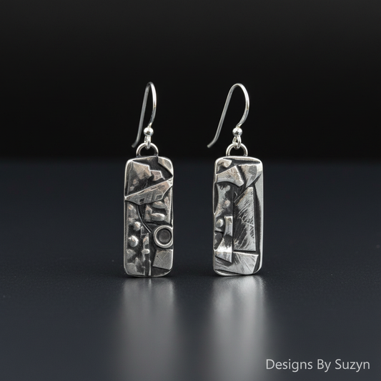 Studio photo of hand-etched Argentium silver botanical dangle earrings standing upright on dark grey reflective surface